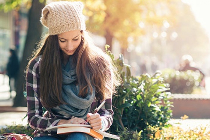 college aged woman sitting outside writing on paper on top of textbook
